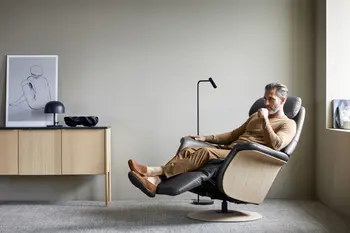 Ein Mann entspannt sich in einem Stressless Sam Sessel mit Holzseiten in der Farbe Paloma Rock und einer runden Basis aus Eiche. Im Hintergrund ein minimalistisches Sideboard mit Deko und eine moderne Leselampe.